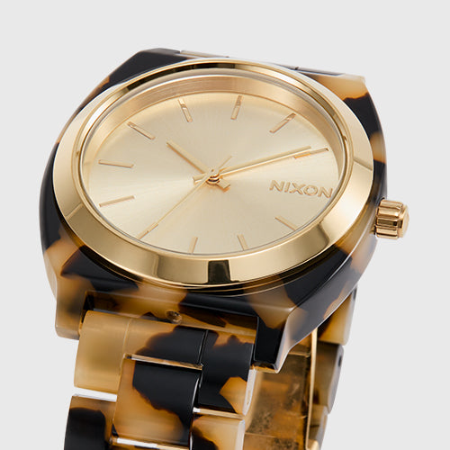 A Nixon analog watch with a gold face and a tortoiseshell-patterned band is displayed against a light background.
